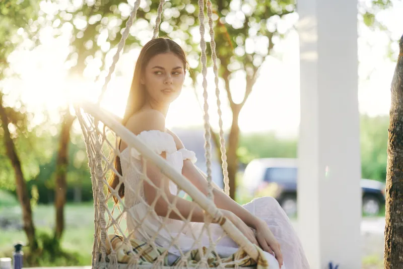 Young woman in a white dress sitting on a swing in a sunlit garden, exuding a serene and contemplative emotion in a natural setting