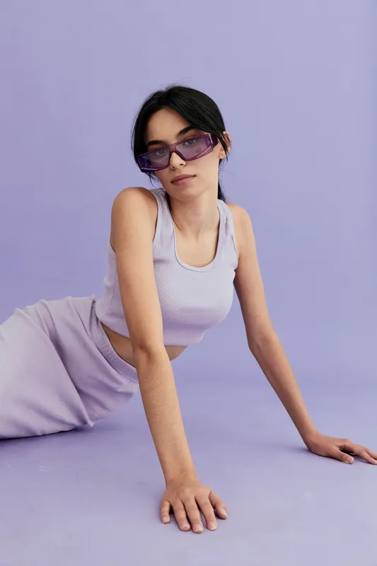 Young woman wearing purple sunglasses and a sleeveless top poses on a lavender background, expressing a modern and relaxed mood with minimalistic style.