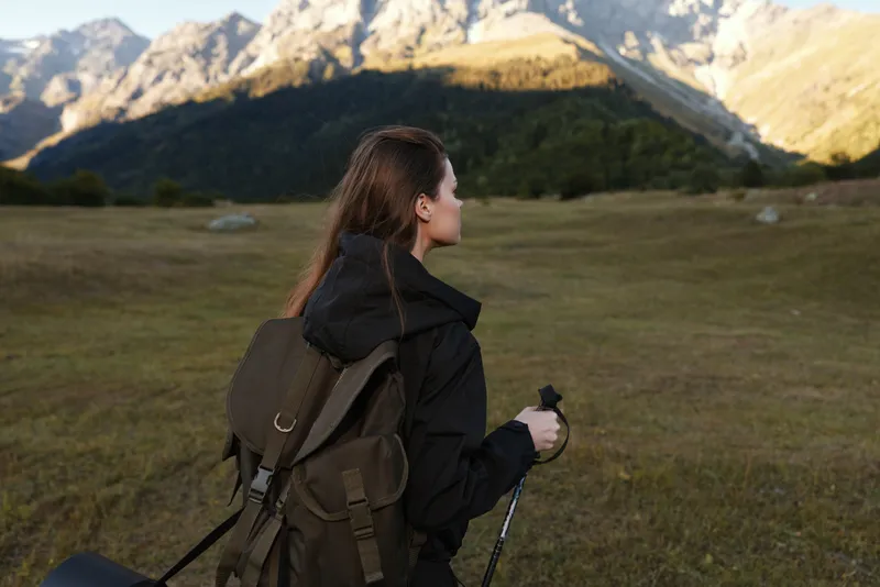 A lone traveler with a backpack admires a scenic landscape in a vast valley. She holds trekking poles and gazes toward distant peaks, embodying exploration, calm focus, and outdoor adventure in open