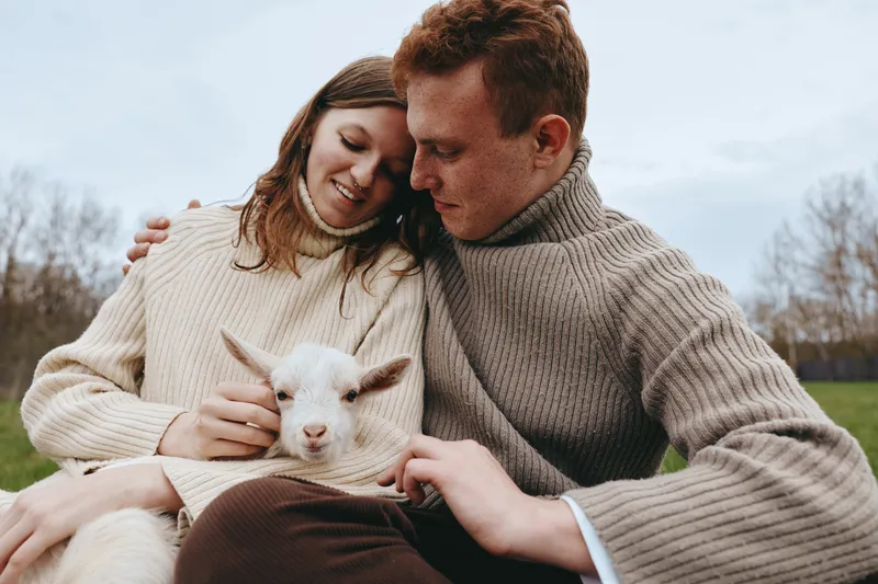 Couple outdoors shares a tender moment with a young goat kid, gentle touch and warm smiles, cozy sweaters, sunlit field, soft breeze, natural tones, peaceful lifestyle scene, rustic charm, animal