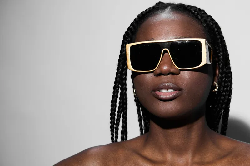 Stylish African model with thick braids sporting oversized sunglasses against a minimalistic gray background, radiating confidence and elegance