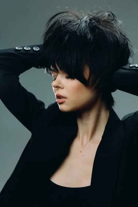 Stylish woman with short black hair posing confidently in a dark outfit against a neutral background.