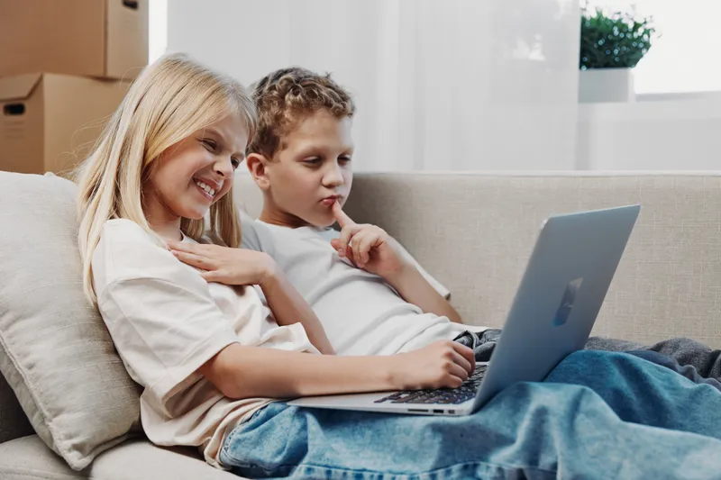 Two children relax on a couch using a laptop together, smiling and focused. Bright living room setting with natural light, showing modern technology and sibling bonding moment.