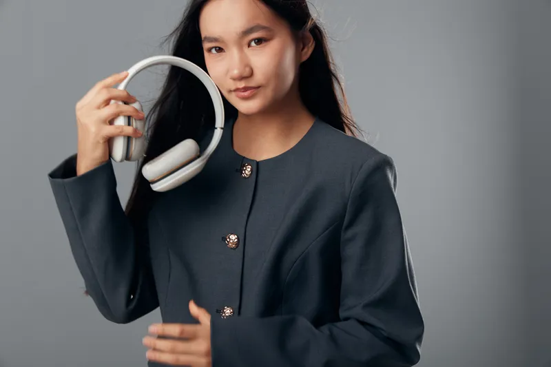 Young woman holding white headphones against gray background, wearing dark jacket with decorative buttons, looking confident and directly at camera in studio portrait.