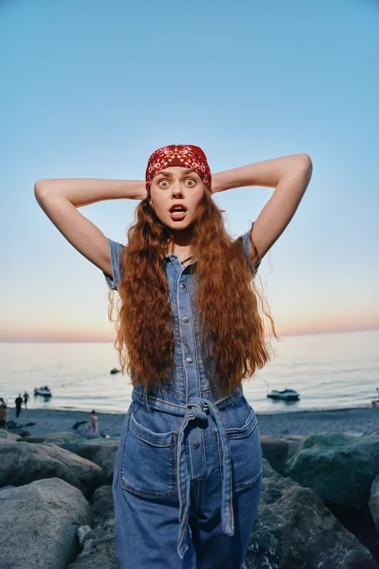 lifestyle woman in modern boho-western denim street style standing on rocks by the sea during sunset with film color effect and surprised expression