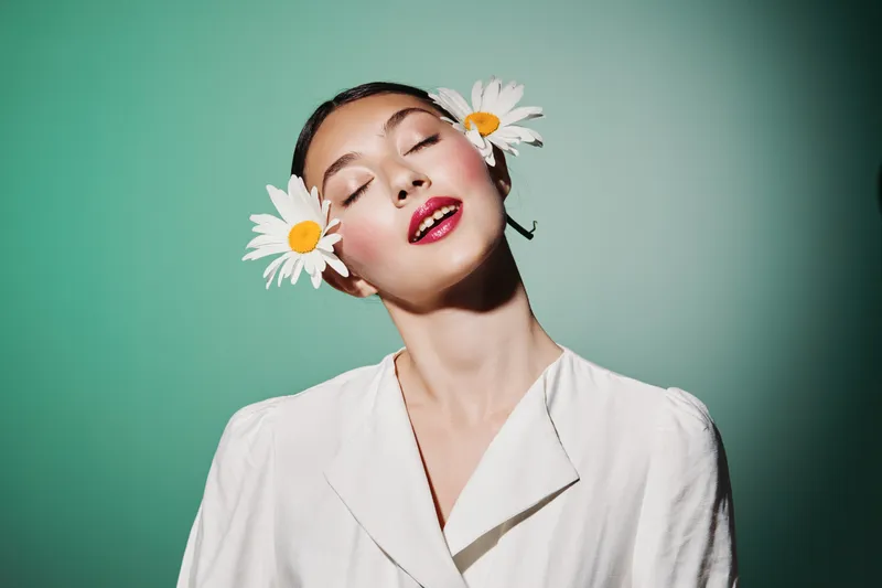 Young woman with daisy flowers in her hair poses with eyes closed wearing a white blazer. Studio shot on green background. Concept of beauty fashion creativity.