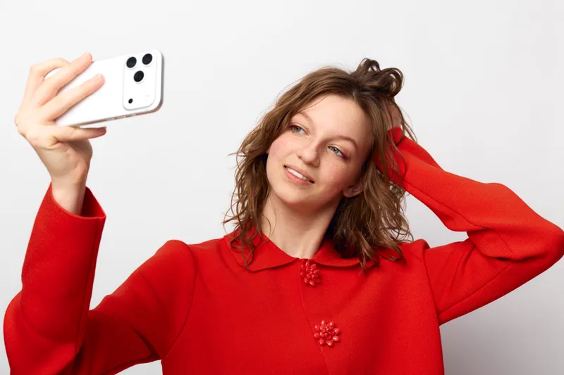 Young woman in red sweater takes selfie with smartphone, smiling and touching hair. White background, casual style, concept of social media, communication, and modern lifestyle.