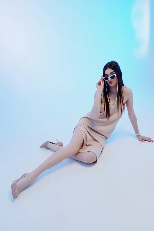 Fashionable woman in a beige dress and sunglasses poses sitting on the floor against a light blue background. Concept of beauty, fashion, elegance, and modern style.