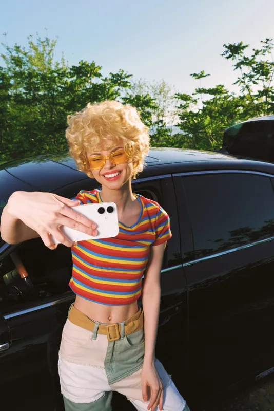 Young woman with blonde curly hair wears a striped crop top and denim shorts, posing against a dark car in bright sun, capturing a carefree selfie moment