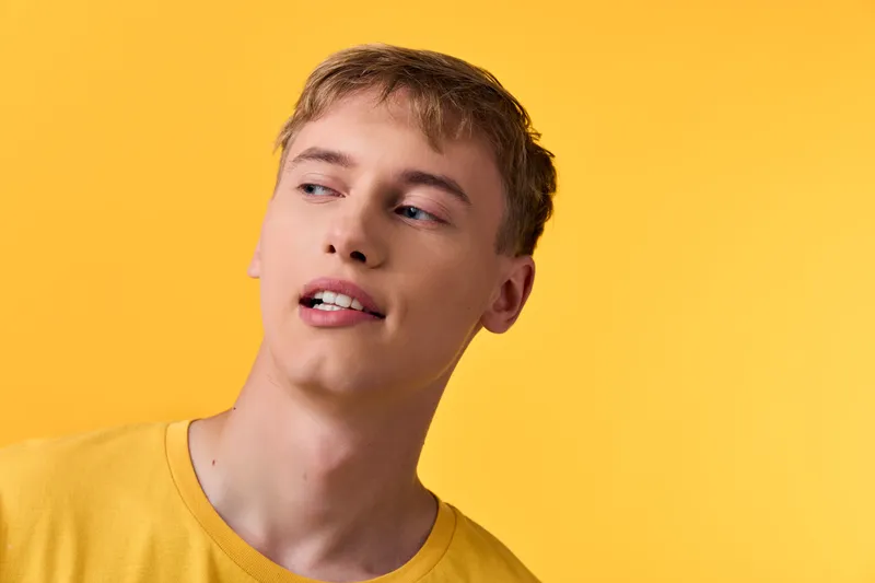 Close up of a male with a relaxed smile against a bright yellow background, wearing a casual yellow tee and looking to the side.