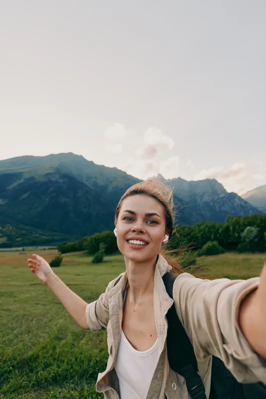 Smiling woman outdoors taking a selfie in a green field with mountains in the background, casual clothing, lifestyle and travel mood.