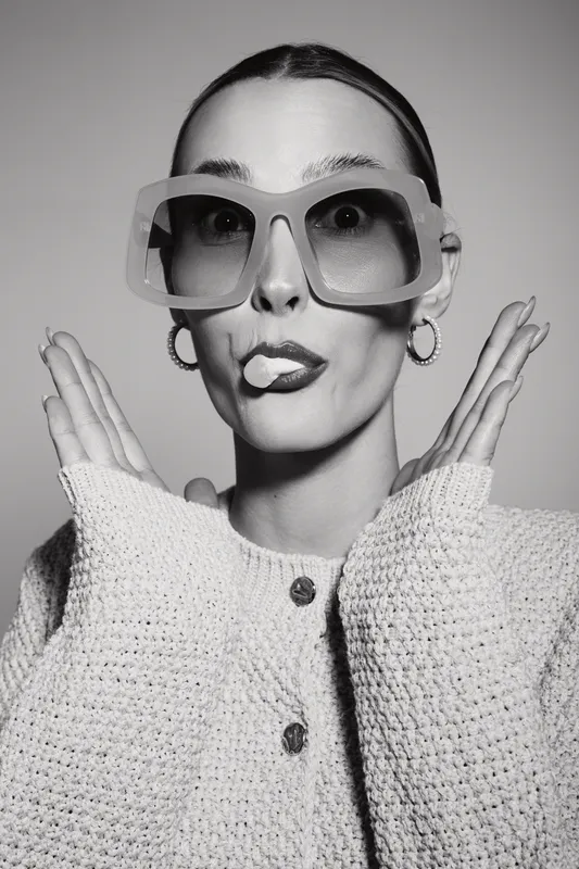 Young Caucasian woman with pale skin and slicked hair makes playful face wearing large sunglasses and knitted sweater on white background.