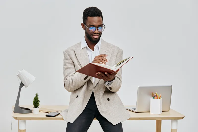 Professional young man with glasses in smart casual attire reading a notebook while standing at a modern desk, workspace setup, indoors, relaxed concentration, professional lifestyle concept.