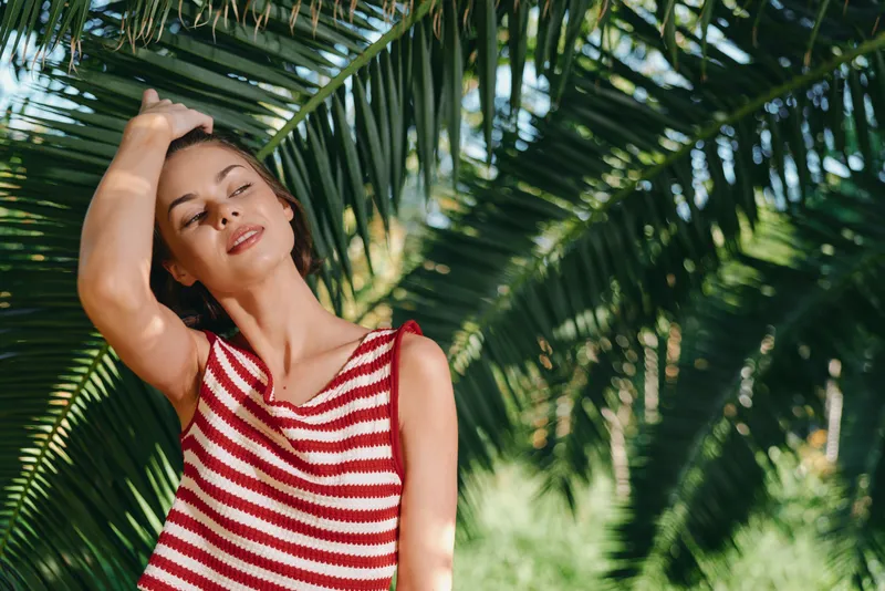 A relaxed woman wearing a red and white striped sleeveless top stands under lush palm leaves in a sunny tropical setting, exuding calm confidence and a breezy, carefree mood.