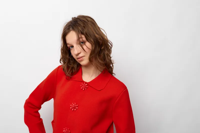 Young woman with curly brown hair wearing a red textured coat poses against a white background. The image conveys fashion, style, and confident casual portrait concept.