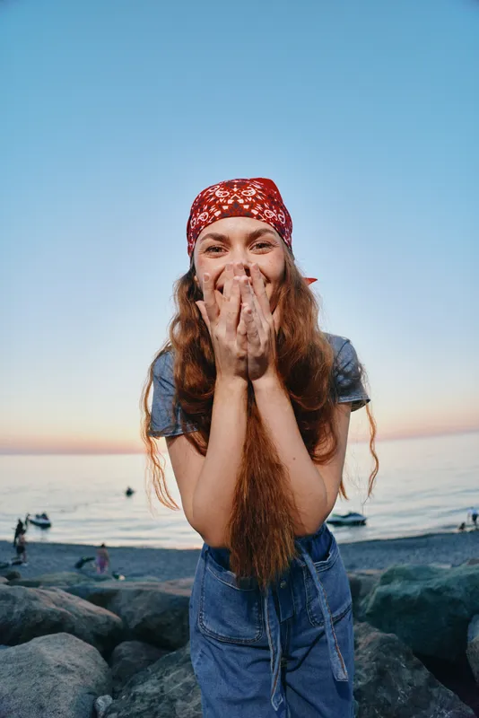 smiling woman wearing modern boho-western denim street style with a red bandana, laughing freely by the rocky seaside, captured in warm film color tones