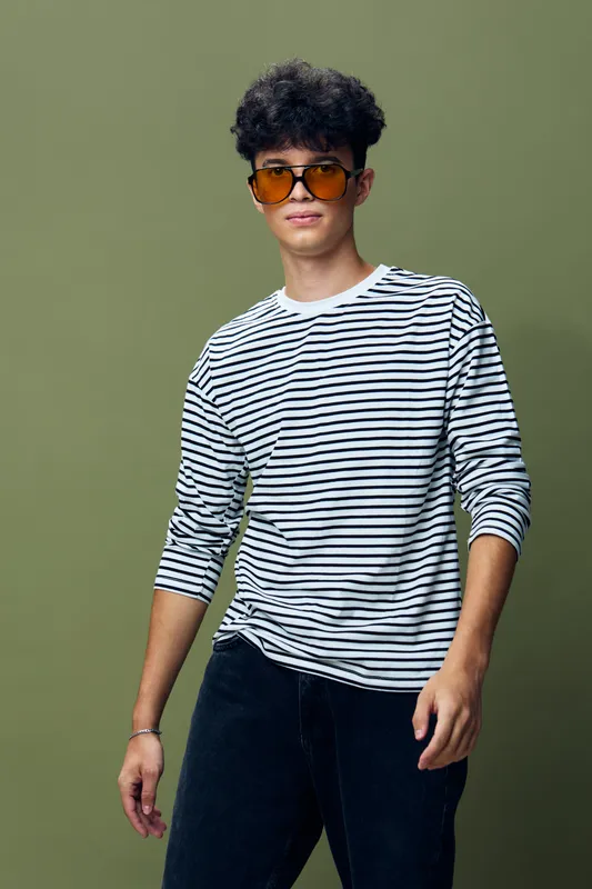 young man wearing striped long sleeve shirt and sunglasses stands confidently against plain olive background, posing casually with relaxed expression and natural hairstyle