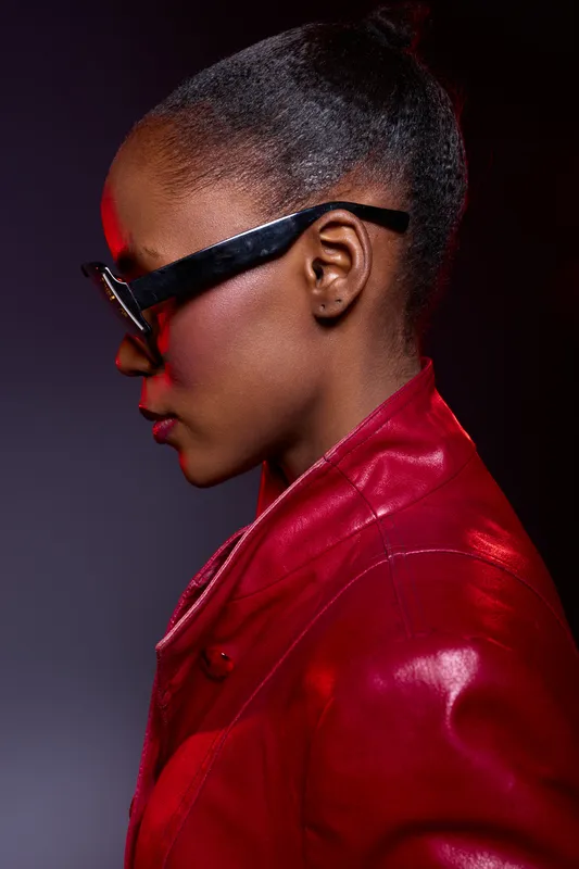 Profile of a young African woman with dark skin wearing black glasses and a red leather jacket. Studio portrait with dramatic lighting, emphasizing fashion and modern style concepts.