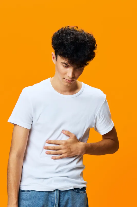 young man with stomach pain wearing casual white t-shirt and blue jeans standing against bright orange background, expressing discomfort and concern