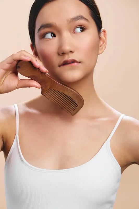 Asian woman with dark hair in a white tank top gently holds a wooden comb to her hair, looking thoughtful and calm in a soft studio setting with warm, even lighting.