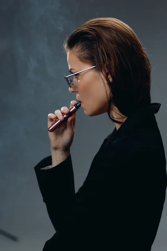 Profile of woman vaping with stylish glasses in dark outfit, smoky background, modern lifestyle concept