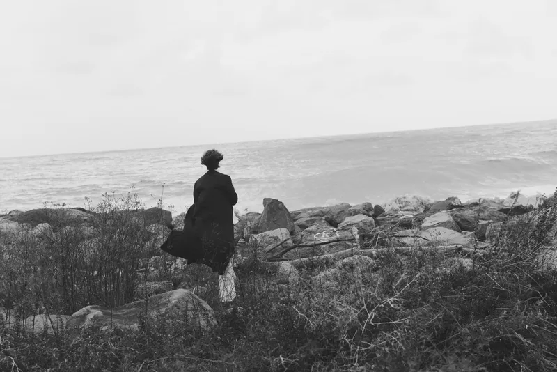 Solitary figure on a rocky coastline gazes toward the sea, a quiet moment of contemplation as the horizon meets the ocean in muted light