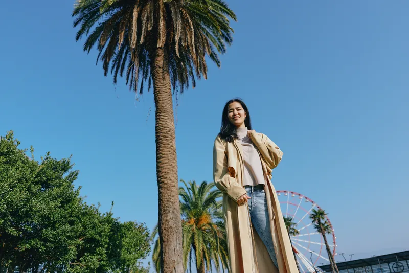 Woman wearing a long beige coat stands among palm trees in a sunny park, with a ferris wheel in the background, casual fashion, outdoor scenery and bright sky.