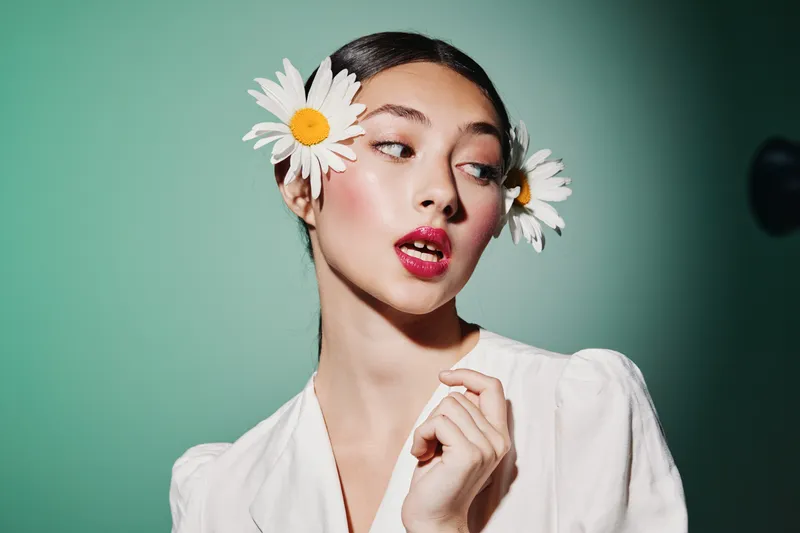 Young woman with daisy flowers in hair wears elegant white blouse, posing against green background. Concept of beauty, fashion, and creative styling studio portrait.