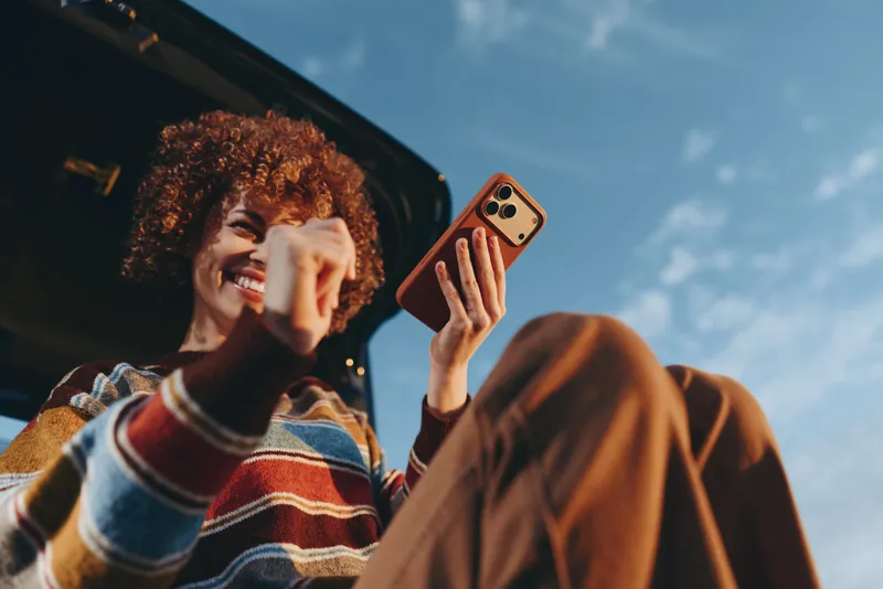 Woman lifestyle scene, outdoors beside car, curly hair woman smiling while talking on phone, warm daylight, casual striped sweater, candid moment for communication and joy
