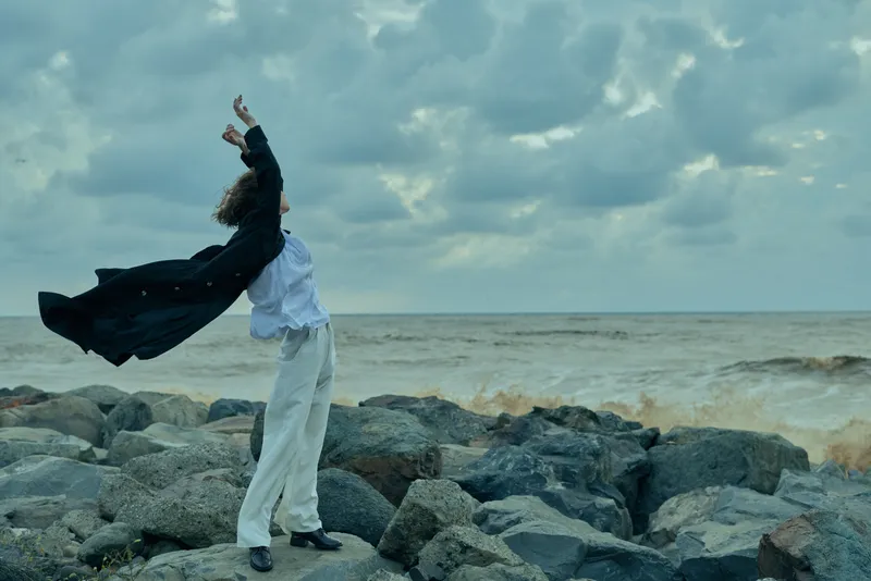 Dynamic figure on a rocky shore with cape billowing, facing the open sea, conveying freedom, mood, and motion against a dramatic, overcast sky that adds cinematic depth