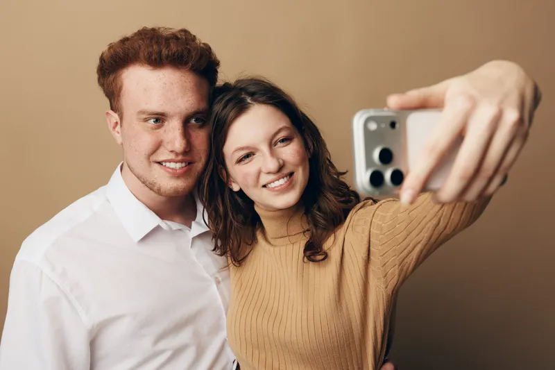 Young couple with red hair and brown hair takes a selfie together smiling. Casual clothing and warm beige background create a cozy and happy atmosphere of friendship and love.