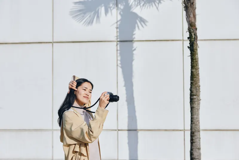Photographer in a beige coat holding a camera outdoors against a white wall with a tall palm shadow, bright daylight, candid and stylish shot highlighting travel, creativity and urban nature,