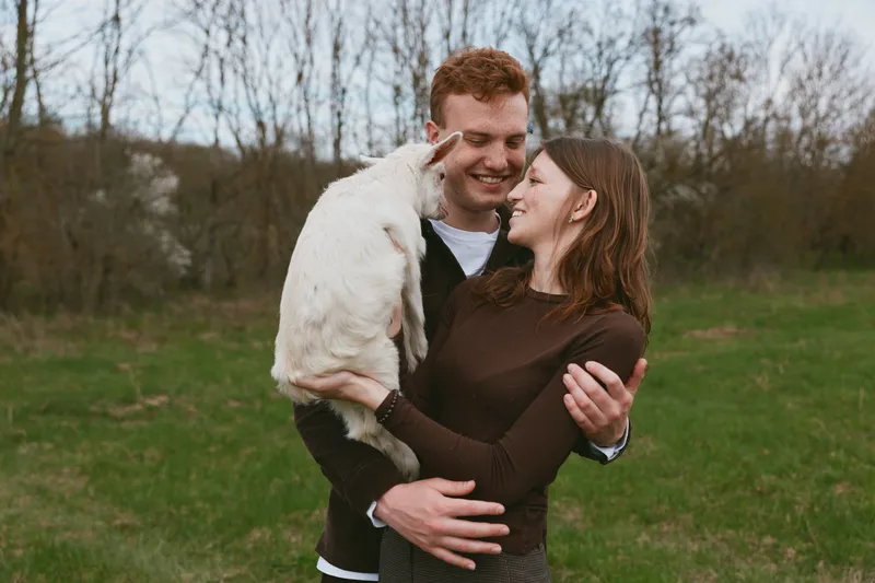 Cheerful couple outdoors with a goat kid, smiling and embracing in a meadow setting, natural light, candid moment, rustic charm, warm colors, joyful connection, countryside lifestyle