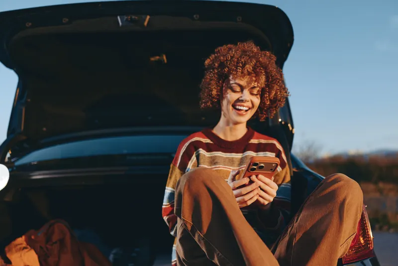 Joyful woman lifestyle in an outdoor car scene, curly hair smiling while holding smartphone, sitting by open trunk, warm sunset light, casual travel mood, candid portrait concept for modern