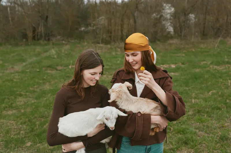 Two women in a countryside field holding goat kids, smiling and interacting with friendly animals. Warm, wholesome outdoor moment focused on care and companionship.