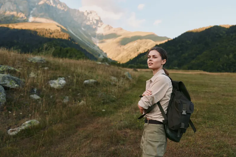 A young woman with a backpack stands in a mountainous landscape with arms crossed, enjoying the outdoor lifestyle and nature adventure during daytime.
