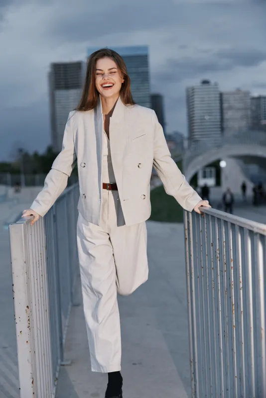 confident woman in light suit posing outdoors smiling with modern city skyline background fashion business style