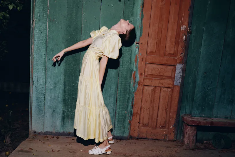 Girl in a pale yellow dress leans back against a weathered wall beside a wooden door at night, creating a mysterious vintage portrait with moody lighting and a quiet, atmospheric tension.