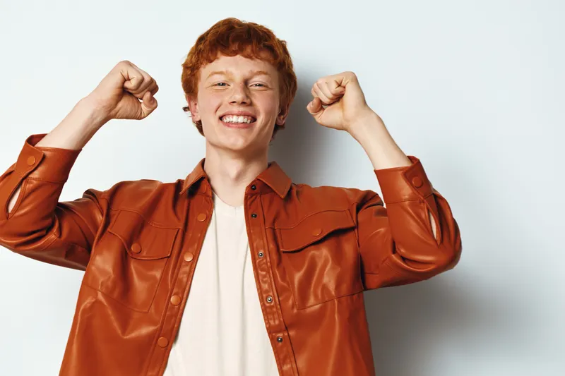 Teenage boy smiling and cheering with raised fists wearing brown leather jacket and beige shirt on white background. Happy youth portrait with positive expression and red hair.