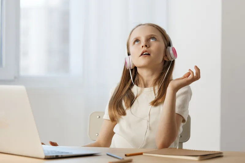 A thoughtful young girl wearing headphones gazes upwards while sitting at a desk with a laptop, reflecting concentration and curiosity in a modern, bright room