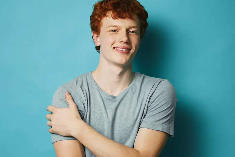 Young man with red hair and casual gray t shirt standing smiling on blue background. Male portrait with confident expression and relaxed posture in studio environment.