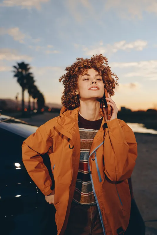 Woman lifestyle on the beach at sunset, curly hair, orange jacket, standing outdoors with calm smile, wind touches hair, warm evening light, soft ocean background, portrait of freedom calm