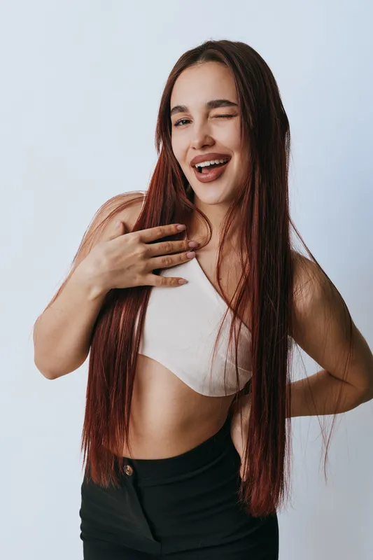 Young woman with long braided hair winking and smiling wearing white crop top and black pants against plain background. Casual confident pose with hand on chest and hip.