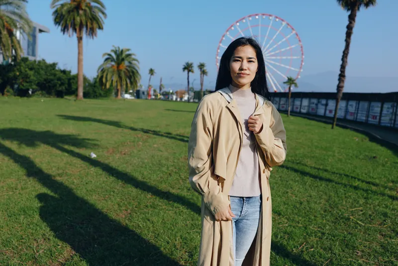 Woman in a beige trench coat stands in a sunny park with a ferris wheel in the background, outdoor scene highlighting casual fashion, leisure, and waterfront ambiance