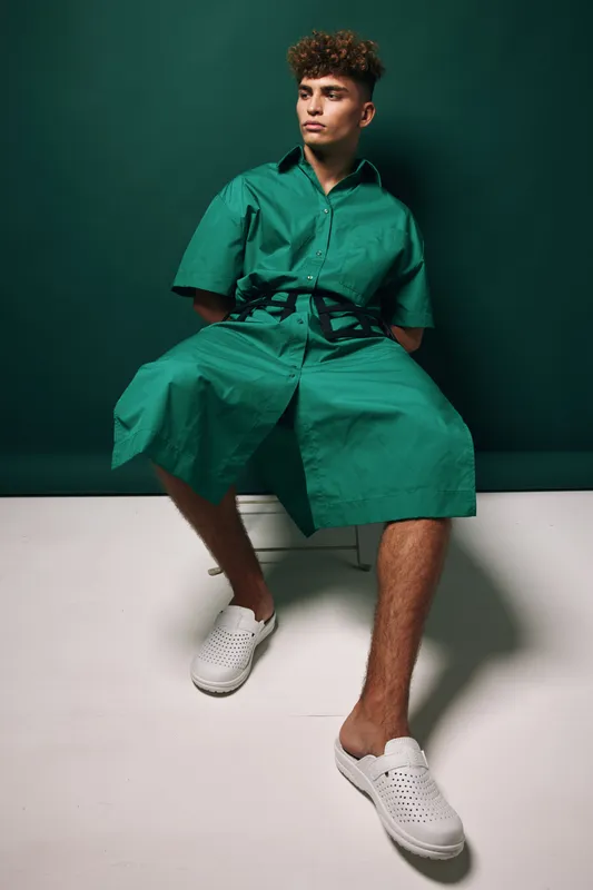 Young man, sitting, posing and wearing green outfit with white shoes in studio. Casual fashion, attitude and trendy with stylish dark curly hair for men modeling apparel and look book.