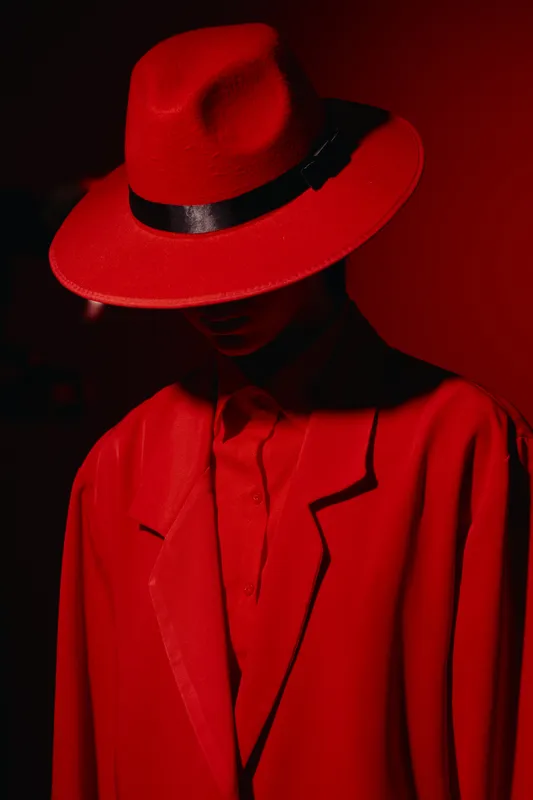 Fashion model wears a vibrant red hat and oversized coat creating dramatic shadows. Creative beauty portrait with strong color contrast and moody lighting on a dark background.