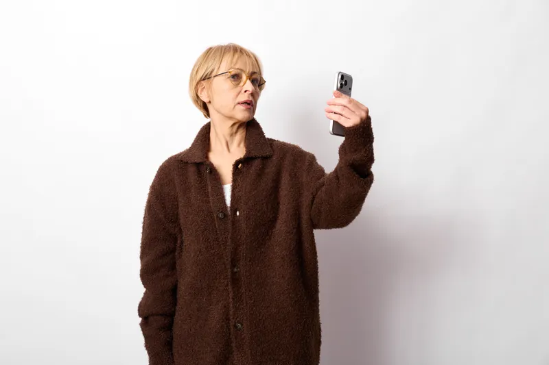 Elderly woman holding smartphone for a selfie, wearing glasses in a studio portrait on white background, technology and communication in indoor setting.