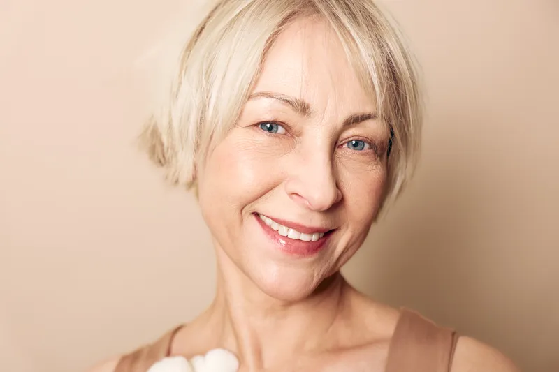 Close-up of smiling mature woman with short blonde hair and blue eyes, holding cotton flowers on beige background. Concept of natural beauty, skincare, wellness, and healthy lifestyle.