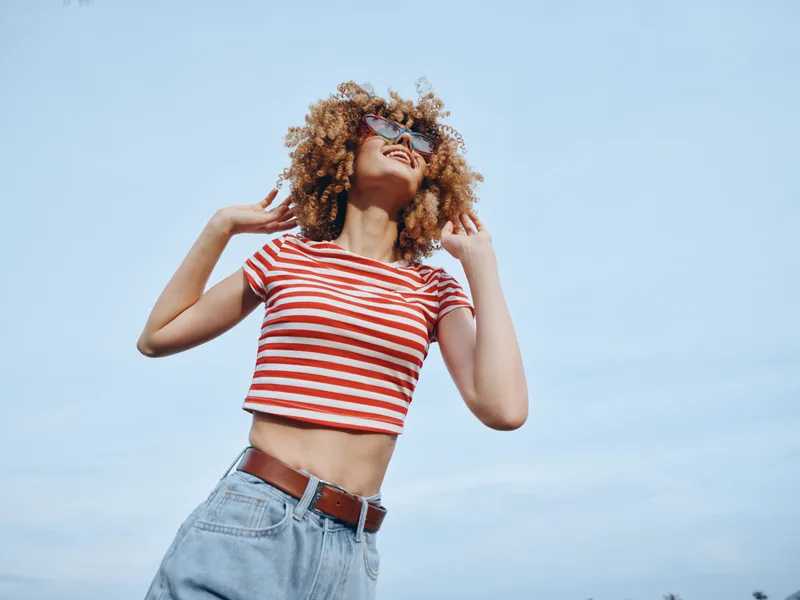 Young woman with curly hair wearing a red and white striped crop top and light denim jeans stands outdoors under a clear blue sky, smiling confidently.