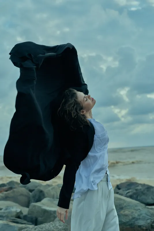Wind sweeps a flowing cape around a woman by a rocky beach, capturing motion and mystery as the ocean meets a cloudy sky, highlighting freedom, drama, and coastal atmosphere in a dynamic moment